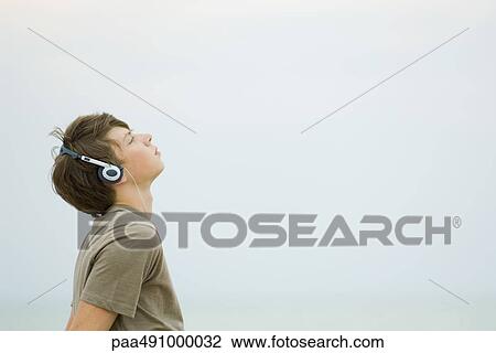Stock Image - Teenage boy listening to headphones, looking up, side view. Fotosearch