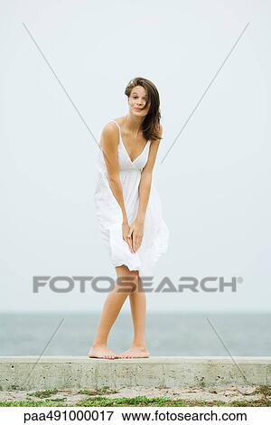 Stock Photo - Teenage girl in front of the sea, bending over to hold skirt, tousled by wind, smiling at camera. Fotosearch