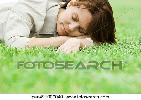 Stock Image - Woman lying on grass with head resting on arms, eyes closed. Fotosearch
