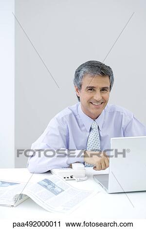 Businessman sitting at desk, smiling at camera View Large Photo Image Stock Image - Businessman sitting at desk, smiling at camera. Fotosearch