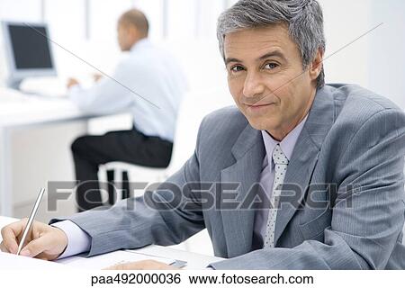 Businessman sitting at desk, using pen, smiling at camera View Large Photo Image Stock Photograph - Businessman sitting at desk, using pen, smiling at camera. Fotosearch