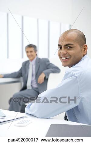 Two businessmen sitting in office, smiling at camera View Large Photo Image Stock Photo - Two businessmen sitting in office, smiling at camera. Fotosearch
