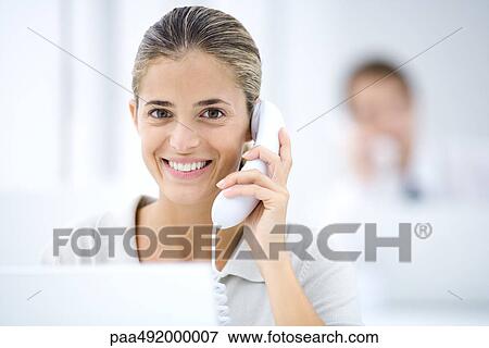Stock Photo - Young woman in office, using landline phone. Fotosearch