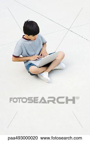 Boy sitting cross-legged on the ground, using laptop View Large Photo Image Stock Image - Boy sitting cross-legged on the ground, using laptop. Fotosearch