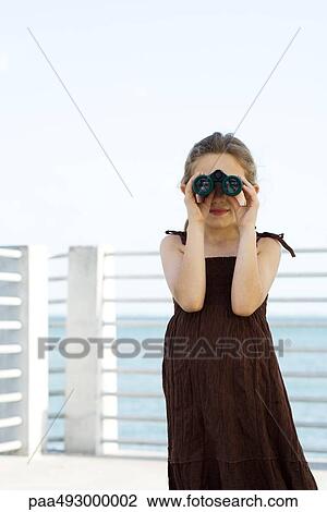 Stock Image - Girl looking through binoculars, standing on pier. Fotosearch