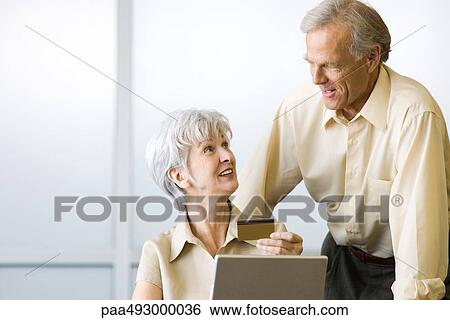 Mature couple looking at each other, woman sitting and holding credit card, man standing View Large Photo Image Stock Photograph - Mature couple looking at each other, woman sitting and holding credit card, man standing. Fotosearch