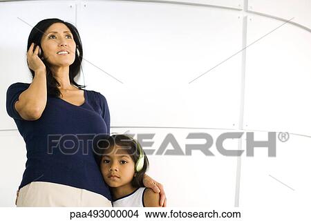 Picture - Mother and daughter standing side by side, listening to headphones, girl looking at camera. Fotosearch