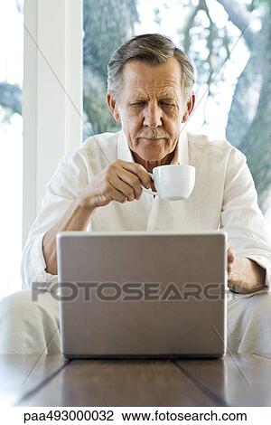 Senior man holding coffee cup, looking down at laptop, sitting with legs apart View Large Photo Image Stock Image - Senior man holding coffee cup, looking down at laptop, sitting with legs apart. Fotosearch