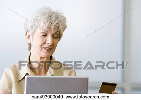 Stock Photo - Senior woman looking at credit card, sitting behind laptop. Fotosearch