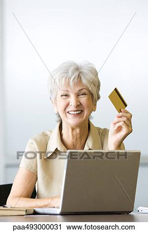 Senior woman sitting at desk, holding credit card, using laptop, smiling at camera View Large Photo Image Stock Image - Senior woman sitting at desk, holding credit card, using laptop, smiling at camera. Fotosearch