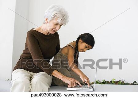 Senior woman sitting behind granddaughter, typing on laptop together View Large Photo Image Stock Photo - Senior woman sitting behind granddaughter, typing on laptop together. Fotosearch