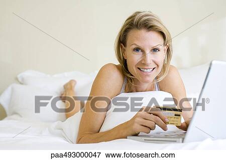 Woman lying in bed, using laptop, holding credit card, smiling at camera View Large Photo Image Stock Photo - Woman lying in bed, using laptop, holding credit card, smiling at camera. Fotosearch