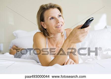 Woman lying on stomach in bed, holding remote control, looking up View Large Photo Image Picture - Woman lying on stomach in bed, holding remote control, looking up. Fotosearch