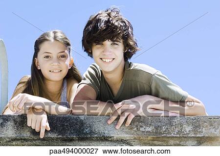 Stock Photo - Brother and sister side by side, leaning on concrete wall, smiling at camera, portrait. Fotosearch