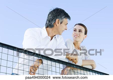 Couple leaning on wire fence, smiling at each other, low angle view View Large Photo Image Picture - Couple leaning on wire fence, smiling at each other, low angle view. Fotosearch