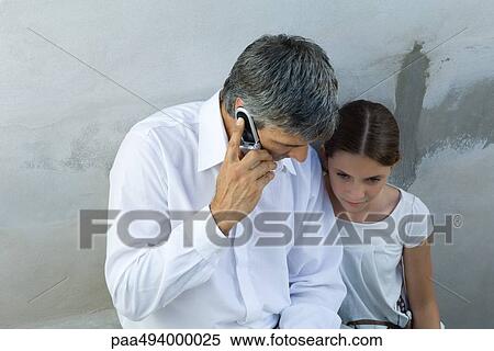 Stock Photography - Father and daughter sitting together, man on cell phone, looking down. Fotosearch
