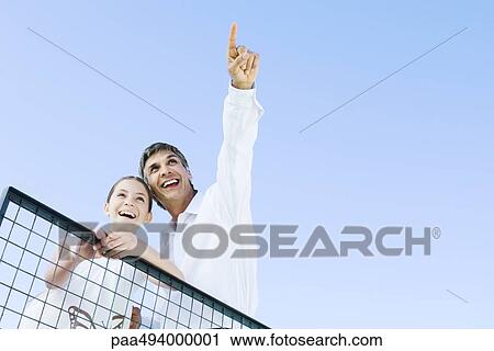 Stock Image - Man standing outdoors with daughter, pointing at the sky, both looking up and smiling, low angle view. Fotosearch