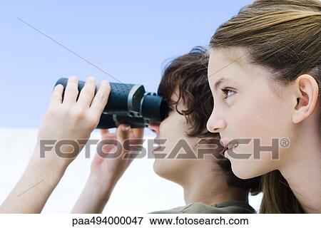 Stock Photo - Teenage boy looking away through binoculars, sister nearby, profile. Fotosearch