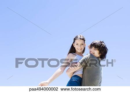 Stock Image - Teenage boy picking up little sister, hugging, low angle view. Fotosearch