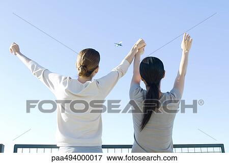 Two women looking at airplane in sky, holding hands, arms raised, rear view View Large Photo Image Stock Photo - Two women looking at airplane in sky, holding hands, arms raised, rear view. Fotosearch