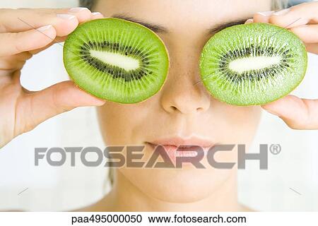 Stock Image - Woman holding kiwi halves in front of her eyes, smiling, headshot. Fotosearch