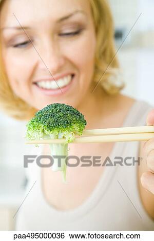 Stock Photograph - Woman using chopsticks to hold a single piece of broccoli, focus on foreground. Fotosearch
