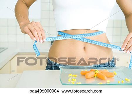 Picture - Woman wrapping a measuring tape around her stomach, plate of vegetables on table. Fotosearch