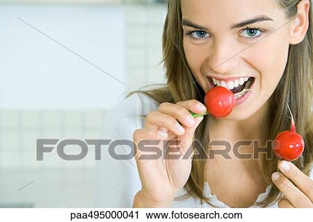 Stock Image - Young woman biting into a radish while holding another, smiling at camera, head and shoulders. Fotosearch
