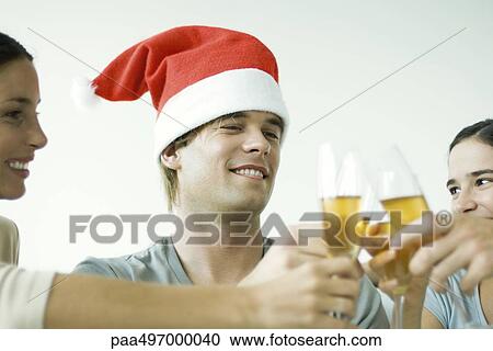 Stock Image - Adult friends clinking champagne glasses, smiling, focus on man in Santa hat, cropped. Fotosearch