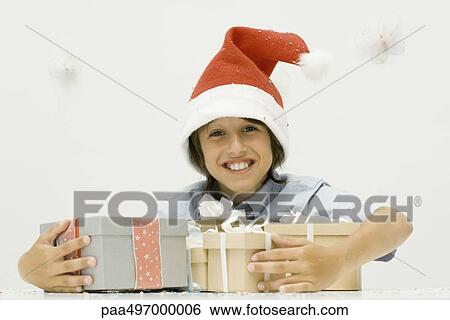 Stock Photograph - Boy holding stack of Christmas gifts, wearing Santa hat, smiling at camera. Fotosearch