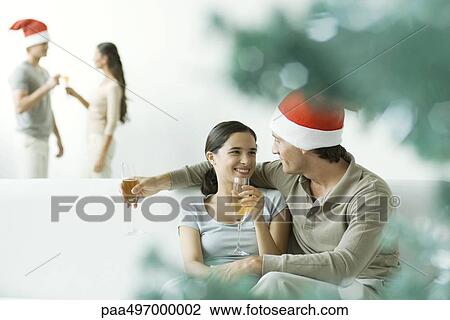 Couple sitting on sofa, drinking champagne, man wearing Santa hat View Large Photo Image Stock Image - Couple sitting on sofa, drinking champagne, man wearing Santa hat. Fotosearch