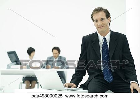 Businessman sitting with laptop, smiling at camera, colleagues in background View Large Photo Image Stock Photography - Businessman sitting with laptop, smiling at camera, colleagues in background. Fotosearch