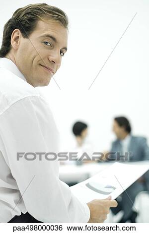 Businessman smiling over shoulder at camera, holding document, colleagues in background View Large Photo Image Stock Photograph - Businessman smiling over shoulder at camera, holding document, colleagues in background. Fotosearch