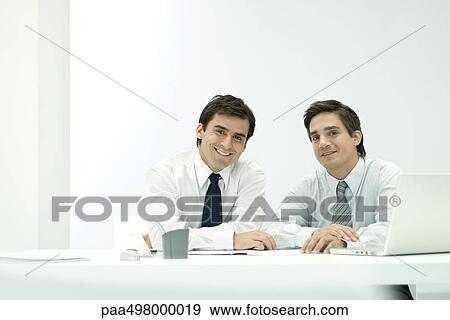 Male business partners sitting side by side at desk, smiling at camera View Large Photo Image Stock Photo - Male business partners sitting side by side at desk, smiling at camera. Fotosearch