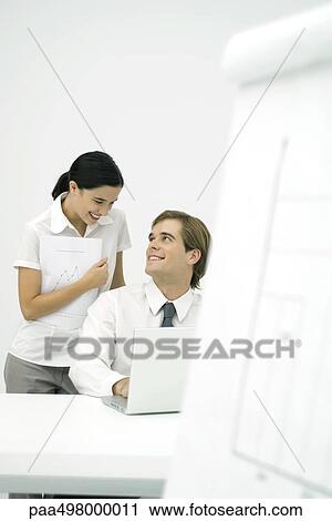 Man sitting at desk, smiling over his shoulder at female colleague standing behind him View Large Photo Image Stock Image - Man sitting at desk, smiling over his shoulder at female colleague standing behind him. Fotosearch