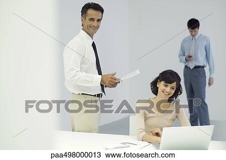Professionals in office, man standing and holding document, woman sitting in front of laptop computer View Large Photo Image Stock Image - Professionals in office, man standing and holding document, woman sitting in front of laptop computer. Fotosearch