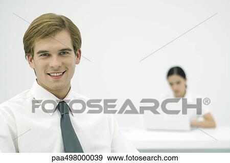 Young professional smiling at camera, colleague in background View Large Photo Image Stock Photo - Young professional smiling at camera, colleague in background. Fotosearch