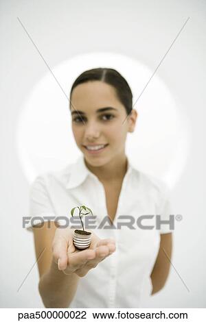 Stock Image - Teen girl holding seedling planted in light bulb base, smiling at camera. Fotosearch