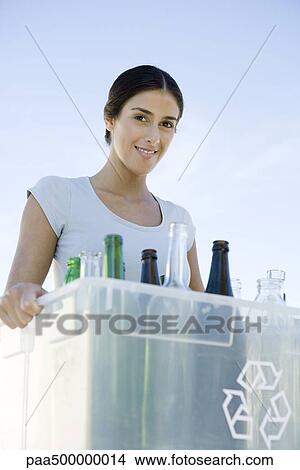 Picture - Woman carrying recycling bin filled with glass bottles, smiling at camera. Fotosearch