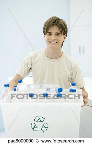 Stock Image - Young man carrying recycling bin filled with plastic bottles, smiling at camera. Fotosearch