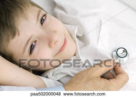 Stock Photo - Doctor holding stethoscope on boy's chest, boy smiling at camera, cropped view. Fotosearch