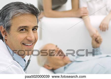 Stock Photo - Doctor smiling at camera, senior man lying on couch in background. Fotosearch