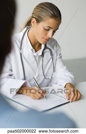 Stock Image - Doctor writing at desk, patient in foreground. Fotosearch