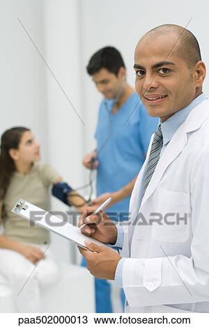 Stock Image - Doctor writing on clipboard, smiling at camera, nurse measuring patient's blood pressure in background. Fotosearch