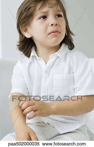 Stock Photography - Little boy picking at adhesive bandage on his arm, frowning. Fotosearch