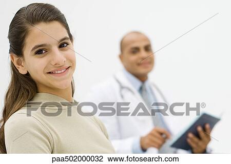 Stock Image - Teenage girl smiling at camera, doctor holding folder in background. Fotosearch