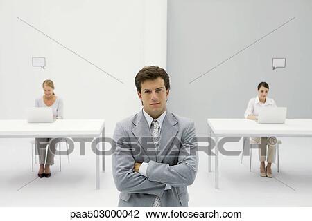 Businessman standing with arms crossed, smirking at camera, women working in background View Large Photo Image Stock Image - Businessman standing with arms crossed, smirking at camera, women working in background. Fotosearch