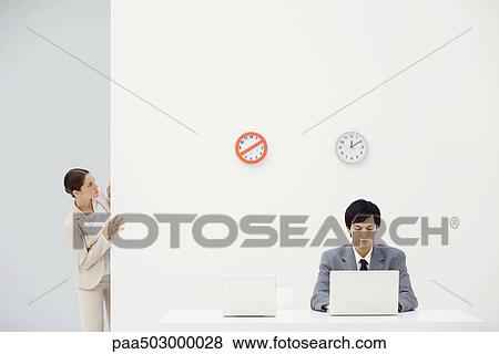 Stock Photo - Female office worker peeking around corner at clocks on wall, colleague looking down at laptop. Fotosearch