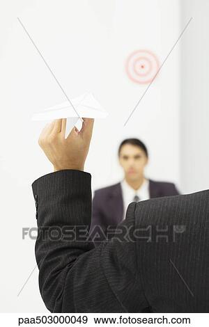 Stock Photo - Man holding paper airplane, aiming at target over colleague's head, cropped view. Fotosearch
