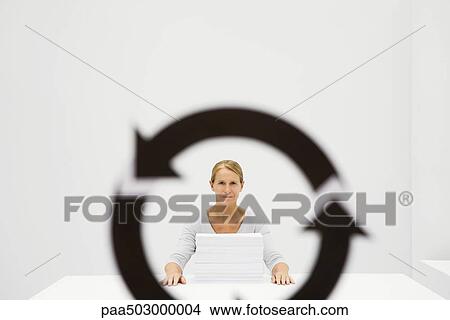 Picture - Professional woman sitting behind stack of paper, looking at camera through recycling symbol. Fotosearch